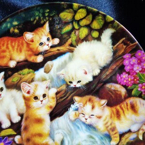 May include: A decorative plate featuring a charming scene of playful kittens. The kittens, in shades of orange and white, are depicted amidst a natural setting with flowers and foliage. The plate's design is detailed and colorful.