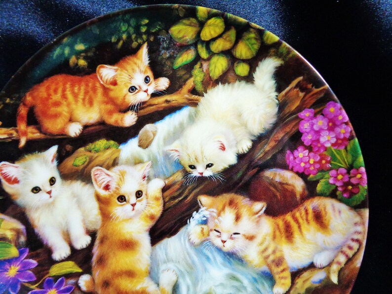 May include: A decorative plate featuring a charming scene of playful kittens. The kittens, in shades of orange and white, are depicted amidst a natural setting with flowers and foliage. The plate's design is detailed and colorful.