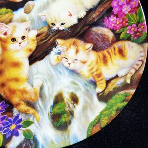 May include: A decorative plate featuring a whimsical scene of playful kittens near a waterfall. The kittens have orange and white fur, surrounded by purple flowers and lush greenery. The plate has a glossy finish.