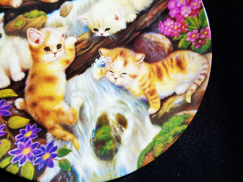 May include: A decorative plate featuring a whimsical scene of playful kittens near a waterfall. The kittens have orange and white fur, surrounded by purple flowers and lush greenery. The plate has a glossy finish.
