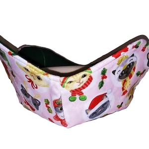 May include: A pink fabric bowl cozy featuring a repeating pattern of cartoon cats and dogs wearing holiday scarves and Santa hats. The interior is a solid dark green. The cozy is designed to hold a bowl of food.