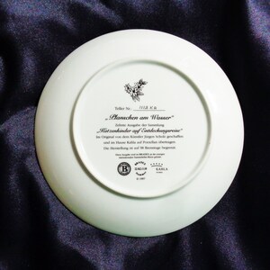 May include: A white porcelain plate with text in German, including "Planschen am Wasser" and "Katzenkinder auf Entdeckungsreise." The plate features a floral design and the Kahla brand logo. The plate is on a dark blue surface.