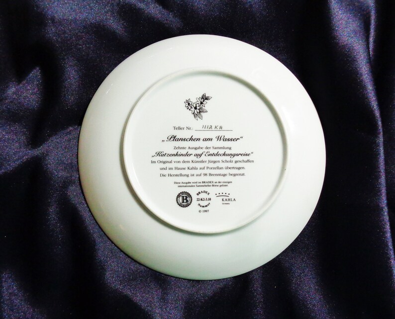 May include: A white porcelain plate with text in German, including "Planschen am Wasser" and "Katzenkinder auf Entdeckungsreise." The plate features a floral design and the Kahla brand logo. The plate is on a dark blue surface.