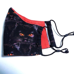 May include: Black face mask featuring a cat design with glowing orange eyes. The mask has a red interior lining and adjustable black ear loops. The cat's face is the focal point of the mask's design.