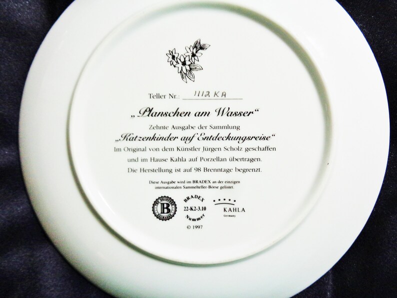May include: White ceramic plate with text in German, including "Planschen am Wasser" and "Katzenkinder auf Entdeckungsreise." Features a floral design and the Bradex and Kahla logos. The plate is a limited edition.