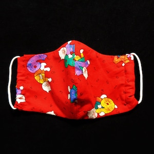 May include: A red face mask with a repeating pattern of colorful numbers and cartoon characters. The mask has white elastic ear loops and is set against a black background.