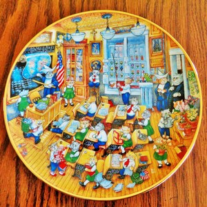 May include: A decorative plate featuring a whimsical school scene with anthropomorphic cats. The cats are depicted as students and teachers in a classroom setting, with desks, books, and an American flag. The plate has a gold rim and is set against a wooden background.