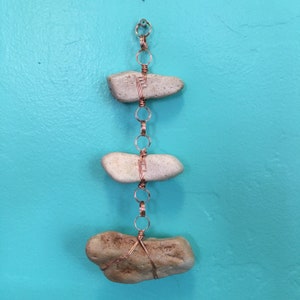 May include: A copper wire chain with three natural stones attached. The stones are different shapes and sizes and are held in place by the wire. The chain is hanging from a hook on a turquoise wall.