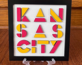 May include: A framed piece of art with the word "KANSAS CITY" spelled out in yellow and pink geometric shapes.