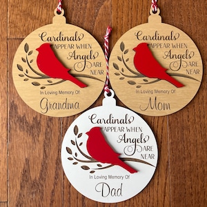 May include: Three wooden Christmas ornaments with a red cardinal bird on a branch. The ornaments are personalized with the words "In Loving Memory Of" and the names "Grandma", "Mom", and "Dad". The ornaments also feature the text "Cardinals Appear When Angels Are Near".