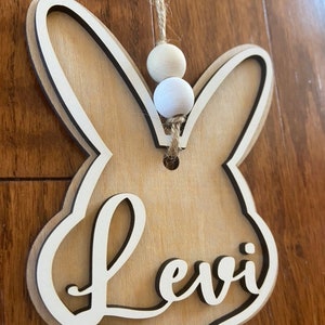 May include: A wooden bunny-shaped ornament with the name "Levi" cut out in the center. The ornament is hanging from a twine string with two white wooden beads.