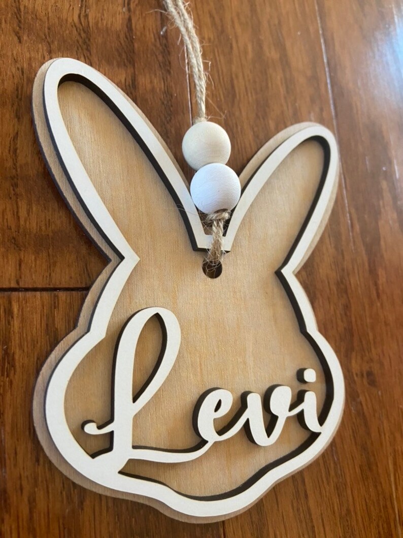May include: A wooden bunny-shaped ornament with the name "Levi" cut out in the center. The ornament is hanging from a twine string with two white wooden beads.
