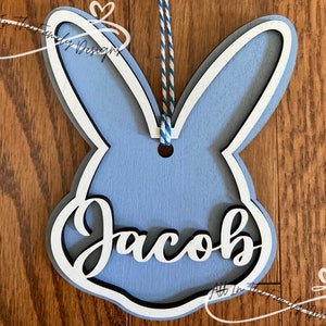 May include: A light blue wooden bunny cutout with a white outline and the name "Jacob" written in white inside. The bunny has a hole at the top for hanging.