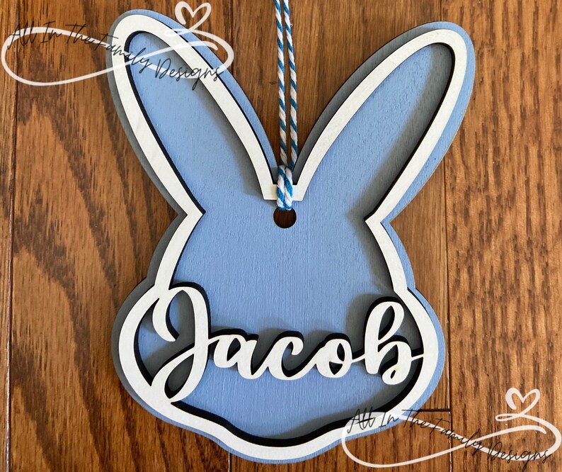 May include: A light blue wooden bunny cutout with a white outline and the name "Jacob" written in white inside. The bunny has a hole at the top for hanging.