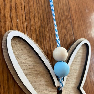May include: Wooden bunny cutout with a white outline and a blue and white striped string with blue and white beads hanging from it.