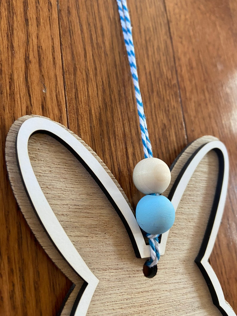 May include: Wooden bunny cutout with a white outline and a blue and white striped string with blue and white beads hanging from it.