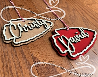 May include: Two layered wooden ornaments in the shape of an arrowhead. One is brown with the name "Christy" in white script, the other is red with "David". Both have white and black borders and red and white twine.