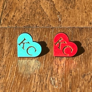 May include: Two wooden heart-shaped ornaments with the letters "KC" carved into them. One ornament is painted turquoise blue and the other is painted red.