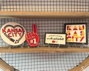 May include: A collection of Kansas City themed decorative items. Includes a heart-shaped sign with "Kansas City" text, a red foam finger with "#1", and two signs with "Kansas City" and "As for me & my house we will watch football 24/7" text.