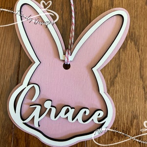 May include: Pink wooden bunny rabbit cutout with a white outline and the name "Grace" written in white with a black outline. The bunny has a hole at the top for hanging.