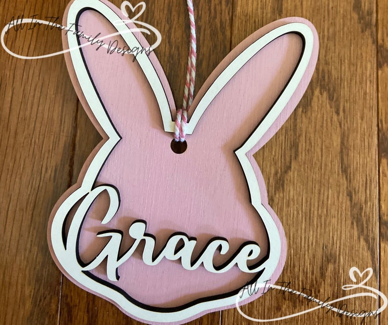 May include: Pink wooden bunny rabbit cutout with a white outline and the name "Grace" written in white with a black outline. The bunny has a hole at the top for hanging.