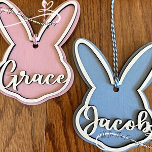 May include: Two wooden bunny shaped ornaments with names "Grace" and "Jacob" painted on them. The bunny on the left is pink and the bunny on the right is blue.