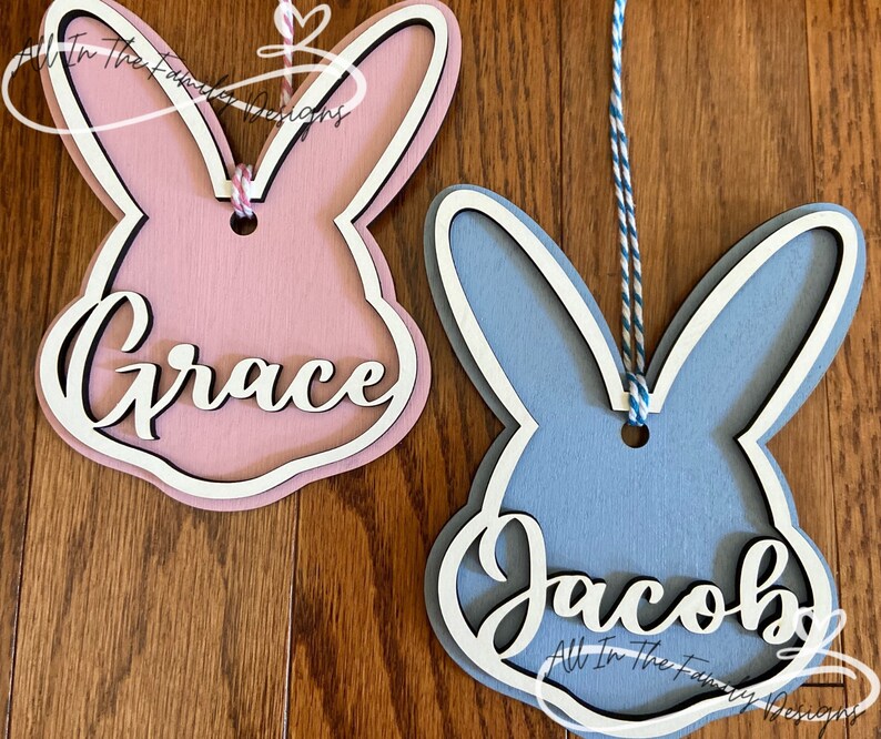 May include: Two wooden bunny shaped ornaments with names "Grace" and "Jacob" painted on them. The bunny on the left is pink and the bunny on the right is blue.