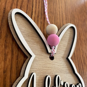 May include: Wooden bunny-shaped door hanger with the name Addison cut out in the center. The hanger is decorated with a pink and white string and wooden beads.