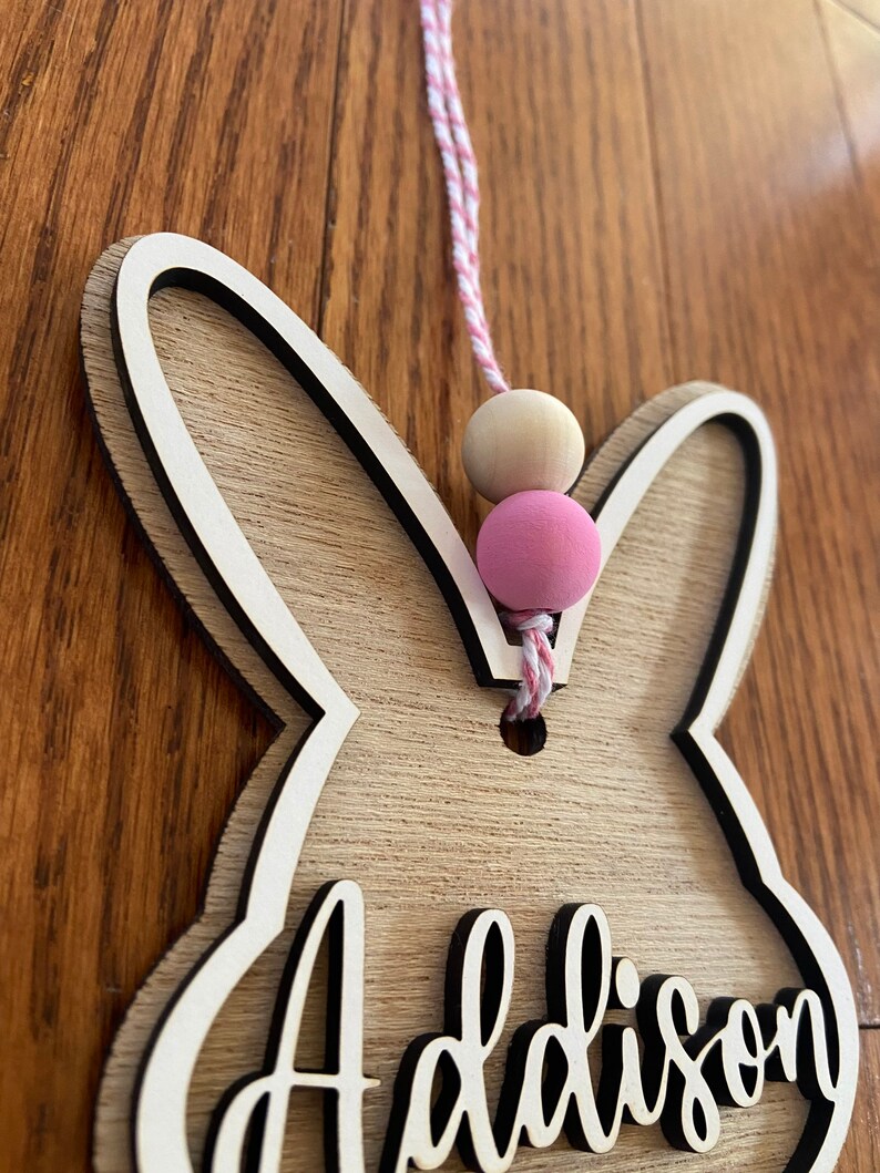 May include: Wooden bunny-shaped door hanger with the name Addison cut out in the center. The hanger is decorated with a pink and white string and wooden beads.