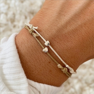 May include: Two friendship bracelets with heart charms. One bracelet is beige and the other is white.