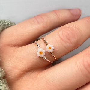 May include: Two delicate rings featuring a white daisy flower charm. The rings are crafted from thin wire and come in silver and gold.