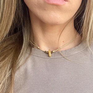 Könnte beinhalten: Eine schlichte, minimalistische Halskette mit einer einzigen, ovalen, hellbraunen Perle. Die Halskette besteht aus einem dünnen, hellbraunen Band und wird von einer Person mit langen, blonden Haaren getragen.