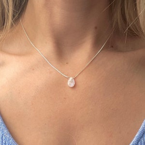 May include: A silver necklace with a small, pear-shaped, pale pink gemstone pendant. The pendant is suspended from a fine silver chain. The necklace is worn over a blue top, creating a simple, elegant look.