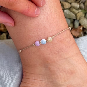 Könnte beinhalten: Ein schlichtes Fußkettchen mit drei kleinen Perlen in den Farben Hellblau, Rosa und Gelb. Das Fußkettchen ist aus einem dünnen, naturfarbenen Band gefertigt.