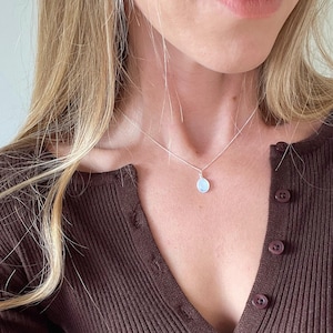 May include: A silver necklace featuring a small, round, pale blue pendant. The pendant is suspended from a fine chain. The person in the image is wearing a brown, ribbed, button-down top.
