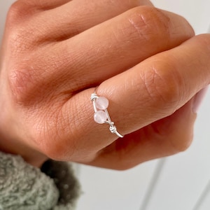 May include: A silver wire ring with two pink beads. The ring is on a person's finger.