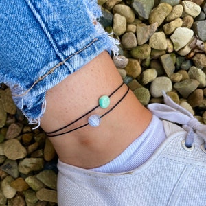 May include: Two anklets with black string and round gemstone beads. One bead is a light green colour and the other is a white and grey colour.