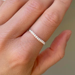 Könnte beinhalten: Ein zarter Silberring, verziert mit einer Reihe kleiner, weißer Perlen. Der Ring ist schlicht und elegant, mit einem minimalistischen Design. Die Perlen sind gleichmäßig angeordnet und verleihen dem Schmuck einen Hauch von Raffinesse.