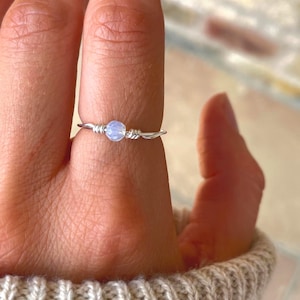 May include: A delicate silver wire ring with a small, round, pale blue bead at the centre. The wire is intricately wrapped around the bead and forms the ring's band. The ring is shown on a finger.