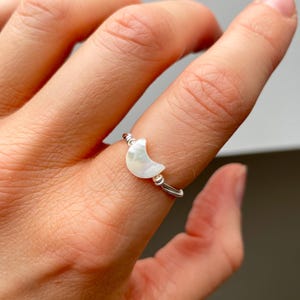 May include: A silver ring with a white crescent moon charm. The ring is adjustable and made of wire.