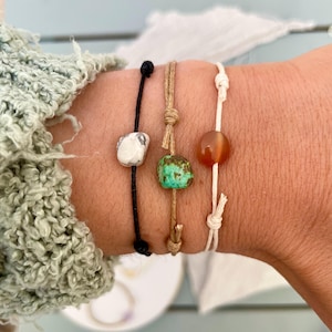 May include: Three handmade bracelets featuring natural stone beads. One has a white and grey stone on a black cord, another a turquoise stone on a tan cord, and the third an orange stone on a white cord.