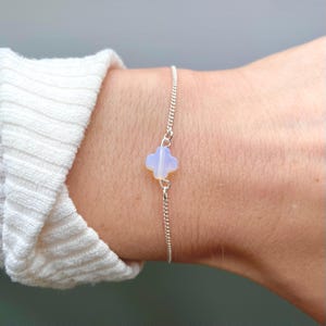 May include: A fine silver chain bracelet with a four-leaf clover charm. The clover is a pale, translucent blue-white colour. The bracelet is worn on a wrist, with a white sweater sleeve in the background.