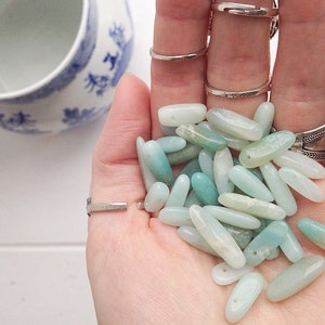 May include: A collection of light green and blue oval-shaped gemstone beads. The beads are smooth and polished, and they are arranged in a pile in the palm of a hand.
