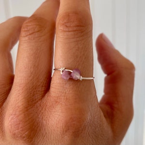 May include: A delicate silver ring with two small, pink, square-shaped stones. The ring is worn on a finger.