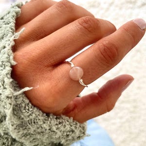 May include: A delicate silver ring with a single pink gemstone. The ring is worn on a finger.
