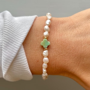 May include: A delicate bracelet made of white pearls with a green cross charm. The bracelet is on a person's wrist.