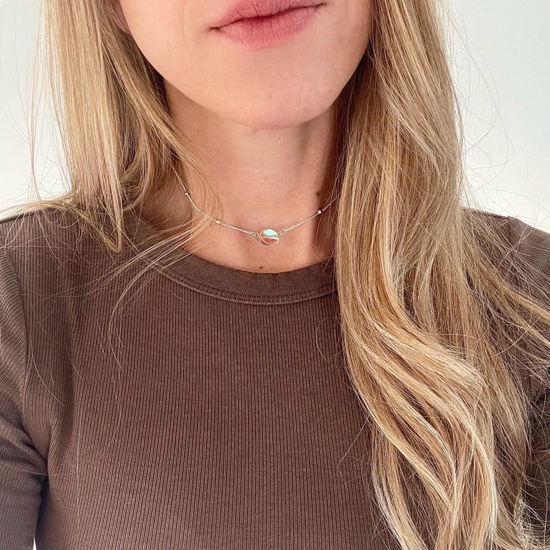 May include: A silver chain necklace with a small, round pendant. The necklace is worn by a person with long, blonde hair and a brown shirt.