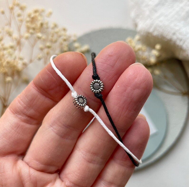 Peut inclure: Deux bracelets d'amiti&eacute;, un blanc et un noir, chacun orn&eacute; d'un petit charm en forme de tournesol argent&eacute;. Les bracelets sont en cordon et pr&eacute;sent&eacute;s sur un fond doux et neutre. Les breloques tournesol ajoutent une touche d&eacute;licate.