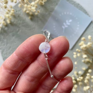 May include: A delicate silver chain bracelet with a round moonstone charm. The moonstone is iridescent and has a milky white color.