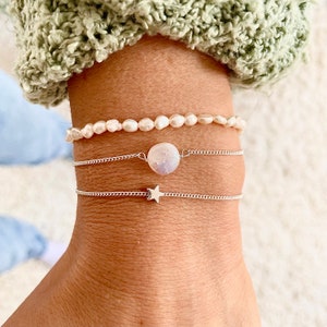 May include: Three bracelets stacked on a wrist. The top bracelet is a pearl strand. The middle bracelet is a silver chain with a large, round, white gemstone. The bottom bracelet is a silver chain with a star charm.